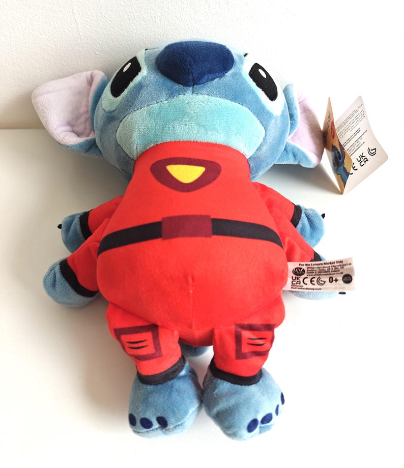 May include: A plush Stitch doll in a red jumpsuit with black accents. The doll has blue fur, large black eyes, and large ears. A yellow triangle is on the chest of the jumpsuit. A tag is attached to the doll.