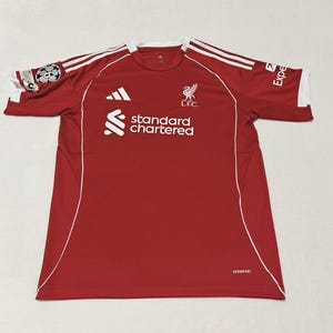 May include: Red soccer jersey with white accents, featuring the Liverpool FC crest and sponsor logos. The jersey has white stripes on the shoulders and the text "standard chartered" on the front.