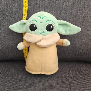 May include: A plush toy of a character with large black eyes, green skin, and large ears. The character is wearing a tan coat with a cream-colored collar. The toy is approximately 10 inches tall.