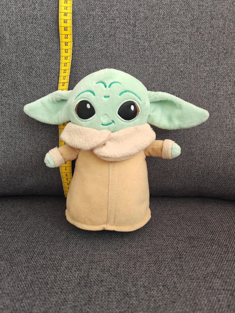 May include: A plush toy of a character with large black eyes, green skin, and large ears. The character is wearing a tan coat with a cream-colored collar. The toy is approximately 10 inches tall.