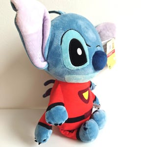 May include: A plush Stitch doll in a seated position. The doll is primarily blue with large ears, a black nose, and a red jumpsuit with black accents and a yellow emblem. The doll has a tag attached to its ear.