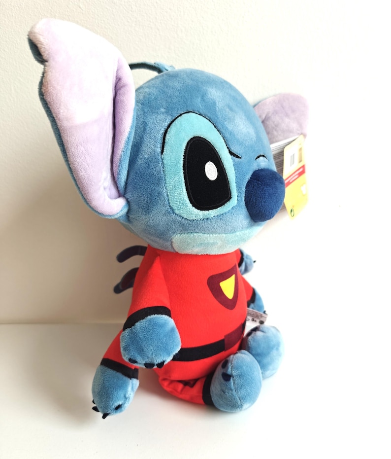 May include: A plush Stitch doll in a seated position. The doll is primarily blue with large ears, a black nose, and a red jumpsuit with black accents and a yellow emblem. The doll has a tag attached to its ear.