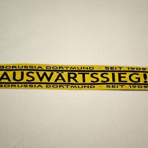 May include: A yellow and black scarf with the text "AUSWÄRTSSIEG!" and "BORUSSIA DORTMUND - SEIT 1909". The scarf has fringed ends and the BVB logo.
