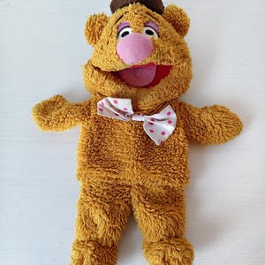 May include: A plush bear toy with golden-brown textured fur, wearing a small brown hat and a white bowtie featuring pink polka dots. The bear has a large pink nose, purple eyelids, and an open mouth showing a red interior and a pink tongue.