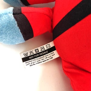 May include: A plush toy with red and black accents, and blue sections. A tag reads "NOT FOR RESALE" with washing instructions and "Made in China". Item No. 71423LD Batch No. 1125 is also printed on the tag.
