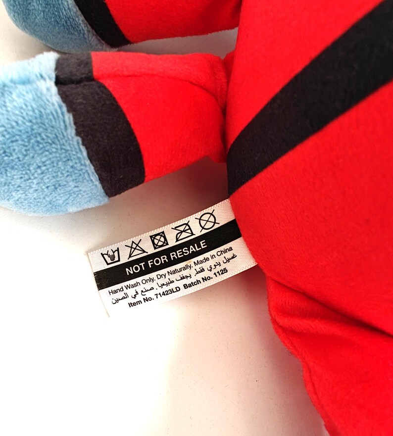 May include: A plush toy with red and black accents, and blue sections. A tag reads "NOT FOR RESALE" with washing instructions and "Made in China". Item No. 71423LD Batch No. 1125 is also printed on the tag.