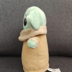 May include: A plush toy of a character from a popular science fiction franchise. The toy has a light green head with large black eyes and large ears. It is wearing a tan coat and has small arms. The toy is sitting on a gray surface.