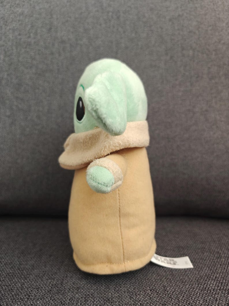 May include: A plush toy of a character from a popular science fiction franchise. The toy has a light green head with large black eyes and large ears. It is wearing a tan coat and has small arms. The toy is sitting on a gray surface.