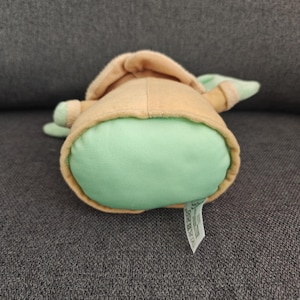 May include: A plush toy of a character from a popular science fiction franchise. The toy has a light green body, tan robe, and large ears. The toy is lying on its back, with a tag visible.
