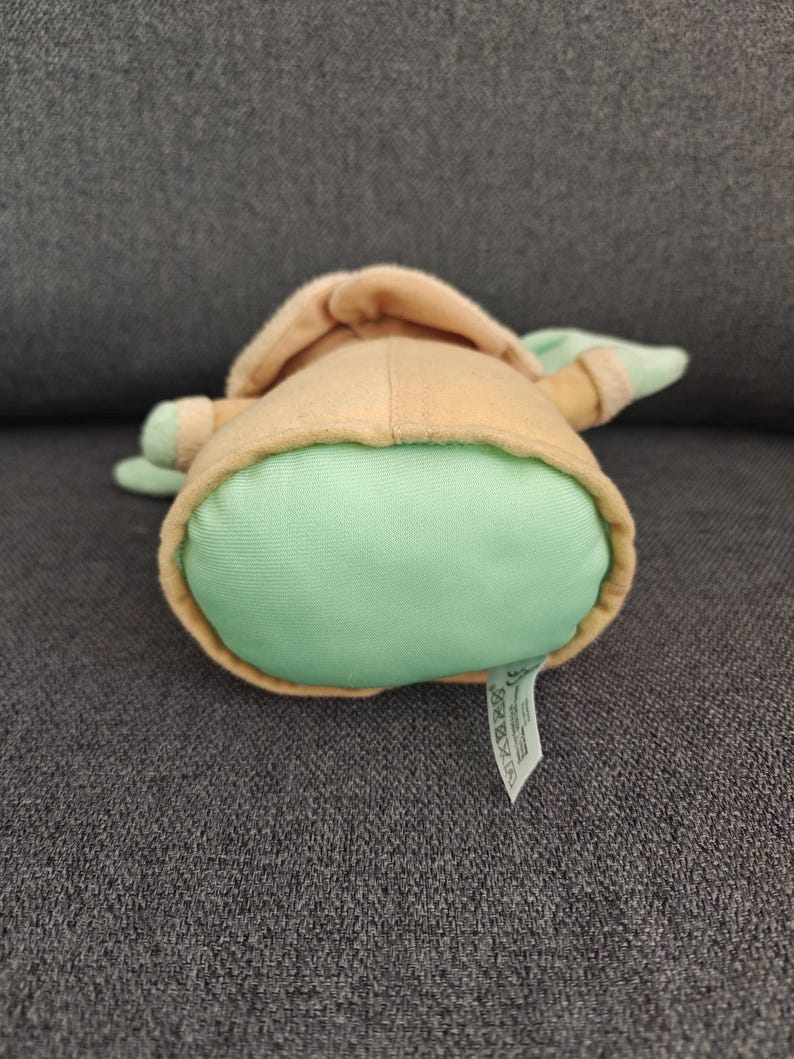 May include: A plush toy of a character from a popular science fiction franchise. The toy has a light green body, tan robe, and large ears. The toy is lying on its back, with a tag visible.