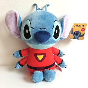 May include: A plush Stitch doll in a red jumpsuit with a yellow triangle on the chest. The doll has large black eyes, blue fur, and large ears. A tag with the Disney Stitch logo is attached.