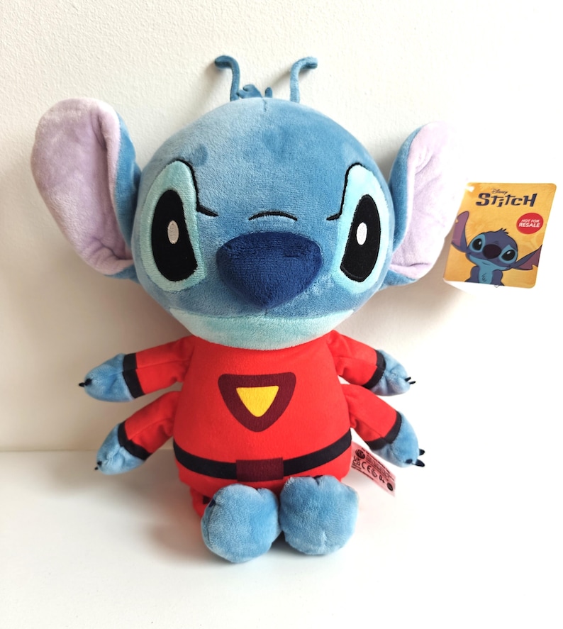 May include: A plush Stitch doll in a red jumpsuit with a yellow triangle on the chest. The doll has large black eyes, blue fur, and large ears. A tag with the Disney Stitch logo is attached.