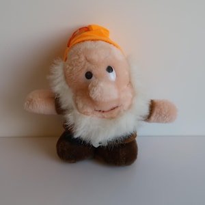 May include: A soft toy dwarf character with a peach-coloured face, large nose, and white beard. The dwarf wears a brown waistcoat, brown shoes, and an orange hat. The toy has a cheerful expression and outstretched arms.