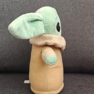 May include: A plush doll of a character with large ears, large black eyes, and a tan robe. The doll has a light green face and ears, with a tan collar and light green accents on the arms.