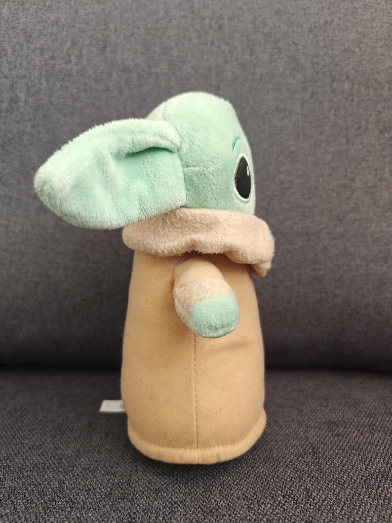 May include: A plush doll of a character with large ears, large black eyes, and a tan robe. The doll has a light green face and ears, with a tan collar and light green accents on the arms.