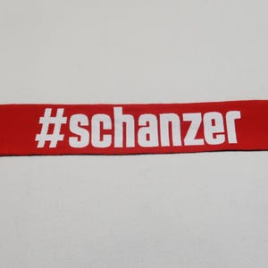 Könnte beinhalten: Rot-schwarzer Fußballschal mit weißem Logo und dem Text "#schanzer".