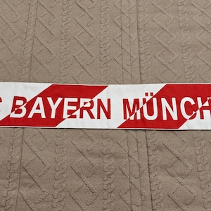 May include: A white and red striped scarf with the text "FC BAYERN MÜNCHEN" in red. The scarf has the Adidas logo and the FC Bayern München team logo. The scarf has white fringe on both ends.