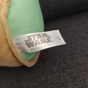 May include: A plush toy featuring a light green head and tan-colored clothing. The Star Wars logo is visible on a white tag. The tag also includes the text "&copy; & TM Lucasfilm Ltd."