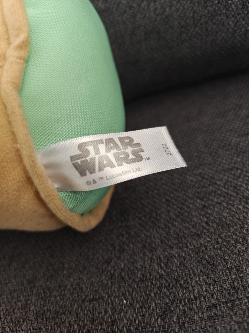 May include: A plush toy featuring a light green head and tan-colored clothing. The Star Wars logo is visible on a white tag. The tag also includes the text "&copy; & TM Lucasfilm Ltd."