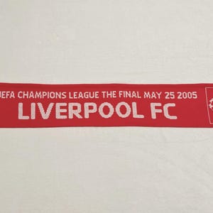 May include: A red scarf with white fringe featuring the Liverpool FC logo and text. The scarf commemorates the UEFA Champions League Final on May 25, 2005. The text "LIVERPOOL FC" is prominently displayed.