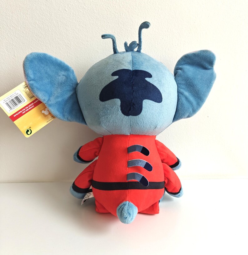 May include: A plush toy of the character Stitch from the Disney movie Lilo & Stitch. The toy has a blue head with large ears, a dark blue patch, and a red body with black accents. The toy is approximately 10 inches tall.