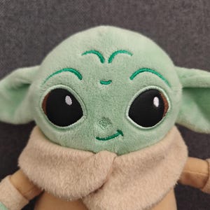 May include: A plush doll of a character with large black eyes, green skin, and large ears. The doll is wearing a tan coat and a beige collar. The doll's face has embroidered details, including a small smile and green markings.