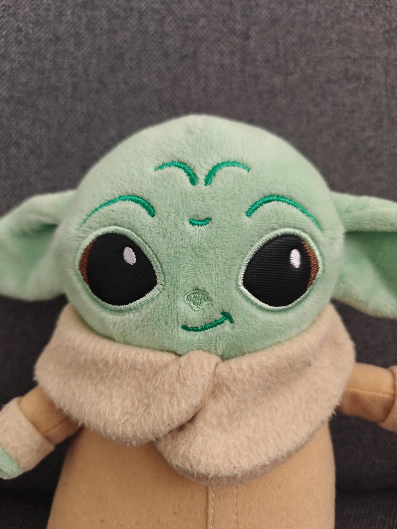 May include: A plush doll of a character with large black eyes, green skin, and large ears. The doll is wearing a tan coat and a beige collar. The doll's face has embroidered details, including a small smile and green markings.