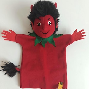 May include: A red devil hand puppet with black hair, red horns, and a green collar. The puppet has red hands and a black tail. A yellow tag with text is attached to the bottom.