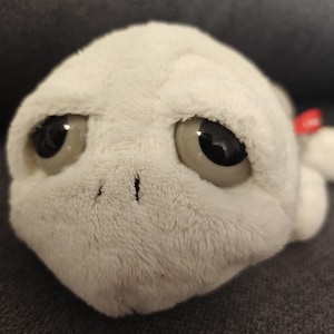 Peut inclure: Une peluche blanche avec de grands yeux noirs. Le jouet a une surface texturée et une petite étiquette rouge. Les yeux sont grands et noirs, donnant au jouet une apparence amicale. Le jouet est posé sur une surface grise foncée.