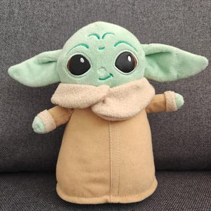 May include: A plush toy of a character with large black eyes, green skin, and large ears. The character is wearing a tan coat with a cream-colored collar. The toy is approximately 10 inches tall.