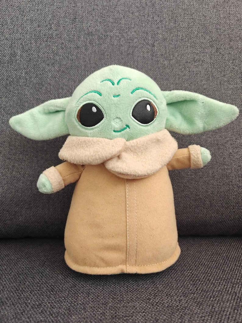 May include: A plush toy of a character with large black eyes, green skin, and large ears. The character is wearing a tan coat with a cream-colored collar. The toy is approximately 10 inches tall.