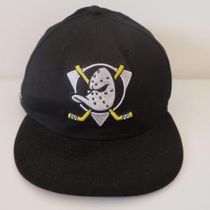 Puede incluir: Gorra de béisbol negra con el logotipo de los Anaheim Ducks. El logotipo incluye una cabeza de pato de dibujos animados, palos de hockey y un triángulo. La gorra tiene una visera curva y una corona estructurada.