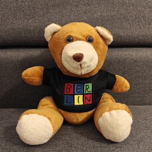Op de afbeelding: Een bruine teddybeer met een zwart t-shirt met het woord "BERLIN" in kleurrijke blokletters. De beer heeft een lichtbruine snuit, crèmekleurige poten en zit op een grijze ondergrond.