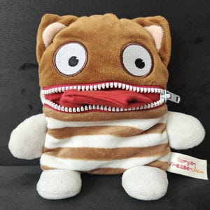 May include: A plush toy with a brown head, large white-rimmed eyes, and a zippered mouth revealing a red interior. The body is brown and white striped, with white arms and feet. The toy has a tag that reads "Sorgen Fresserchen."