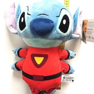 May include: A plush Stitch doll in a red jumpsuit with black accents and a yellow triangle. The doll has blue fur, large black eyes, and large ears. A measuring tape is next to the doll.