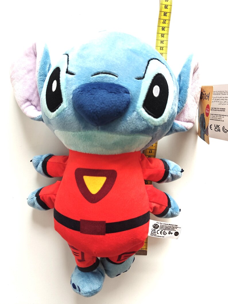 May include: A plush Stitch doll in a red jumpsuit with black accents and a yellow triangle. The doll has blue fur, large black eyes, and large ears. A measuring tape is next to the doll.