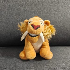 Może przedstawiać: Pluszowa zabawka przedstawiająca tygrysa szablozębnego, z pomarańczowym futrem, dużymi białymi kłami i czerwonym nosem. Zabawka ma przyjazny wyraz twarzy z zielonymi oczami i siedzi na szarej powierzchni.