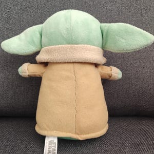 May include: A plush toy of a character from Star Wars, featuring a light green head and large ears, a tan body, and small arms. The toy has a tag that reads "Star Wars."