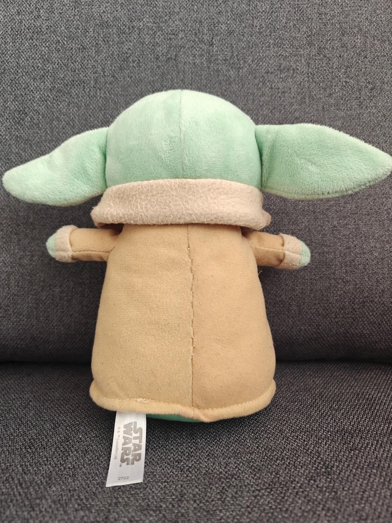 May include: A plush toy of a character from Star Wars, featuring a light green head and large ears, a tan body, and small arms. The toy has a tag that reads "Star Wars."