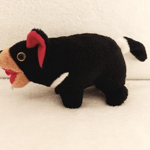 Puede incluir: Un peluche que representa un demonio de Tasmania. El juguete es principalmente negro con una cara marrón, orejas rojas y una franja blanca en el costado y la cola. La boca está abierta, revelando el interior rojo.