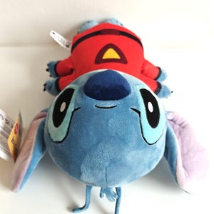 May include: A plush toy featuring the character Stitch from Lilo & Stitch, in a light blue color with large black eyes and long ears. A red plush figure with a yellow triangle is on top of Stitch.