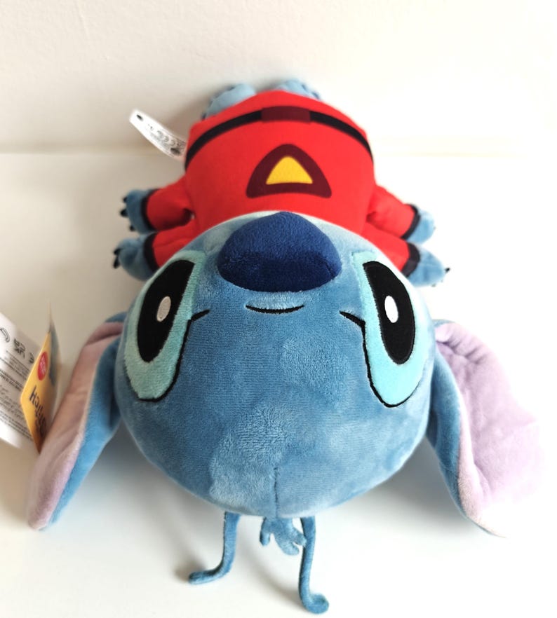 May include: A plush toy featuring the character Stitch from Lilo & Stitch, in a light blue color with large black eyes and long ears. A red plush figure with a yellow triangle is on top of Stitch.