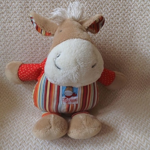 May include: A plush donkey toy with a white face, brown ears, and a tuft of white hair. It wears a red polka-dot shirt and a striped top with the "Sigikid" logo. The toy is soft and cuddly.