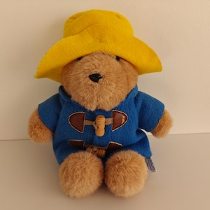 Può includere: Un orsacchiotto di peluche con pelliccia marrone, che indossa un cappello giallo e un cappotto blu con chiusure a toggle in legno. L'orso è seduto. Una piccola etichetta è attaccata alla gamba dell'orso.