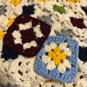 May include: Close-up of two crocheted granny squares in burgundy and blue, with a cream-colored background. The burgundy square has cream-colored accents, while the blue square features a yellow and cream flower design. The background is a cream-colored crocheted blanket.