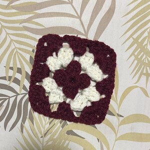 May include: A square, handmade crochet granny square in a floral pattern. The square features a deep burgundy border and center, with white flower-shaped accents. The yarn appears to be a soft, textured material, perfect for crafting projects.