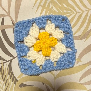 May include: A square, crocheted granny square in shades of blue, cream, and yellow. The center features a yellow flower shape, surrounded by cream-colored petals. The outer border is a light blue, creating a vibrant, handmade textile.
