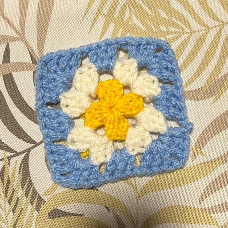 May include: A square, crocheted granny square in shades of blue, cream, and yellow. The center features a yellow flower shape, surrounded by cream-colored petals. The outer border is a light blue, creating a vibrant, handmade textile.