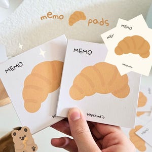 May include: White memo pads featuring a cartoon croissant design. The word "MEMO" is printed on the pads, with "bapstudio" below. Additional pads and a small bear sticker are also visible in the image.
