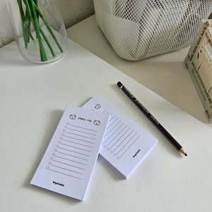 Könnte beinhalten: Zwei weiße Checklisten-Notizblöcke mit einem Bärenkopfdesign und dem Text "check list" oben. Die Notizblöcke liegen auf einer weißen Oberfläche mit einem Bleistift.